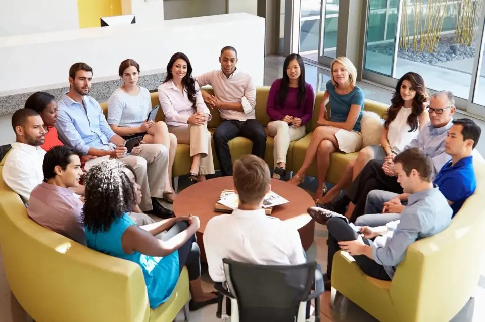 image-5 A diverse group of people sitting in a circle of chairs, engaged in a discussion in a bright, modern room.
